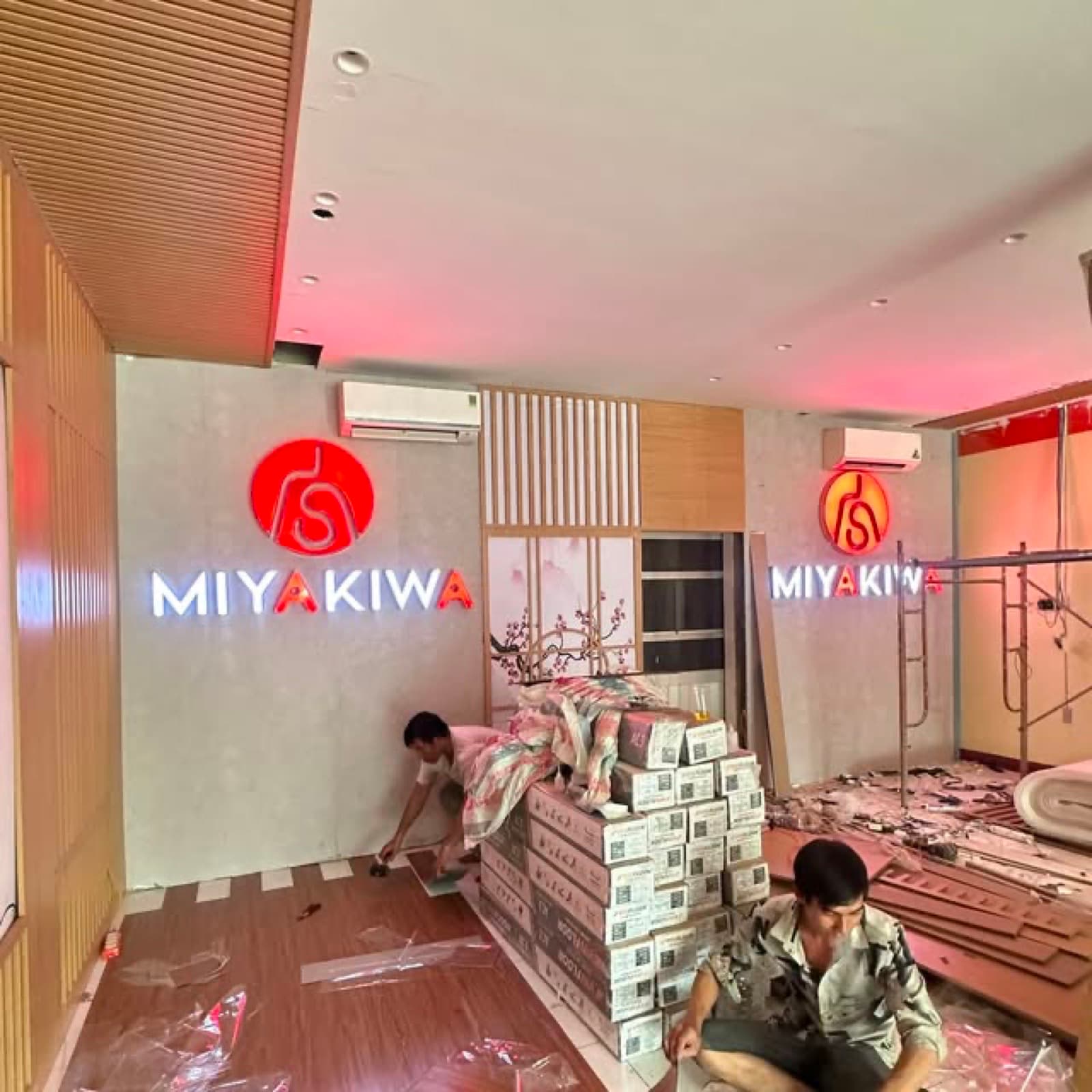 Hai người thợ trong không gian MIYAKIWA có bảng chữ sáng và các kiện vật liệu